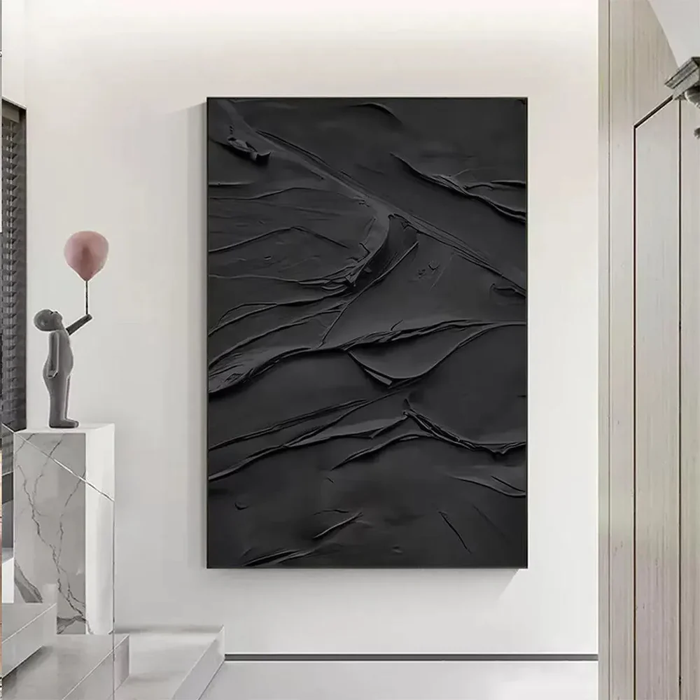 Black textured abstract painting hangs on a white wall, with raised dark strokes beside a simple pedestal and vase in a minimalist interior.