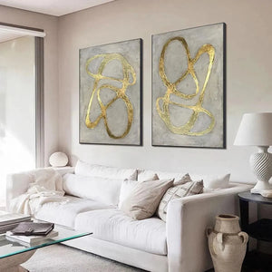 Two vertical abstract paintings with textured gray backgrounds and raised gold lines, black framed, in a living room setting.