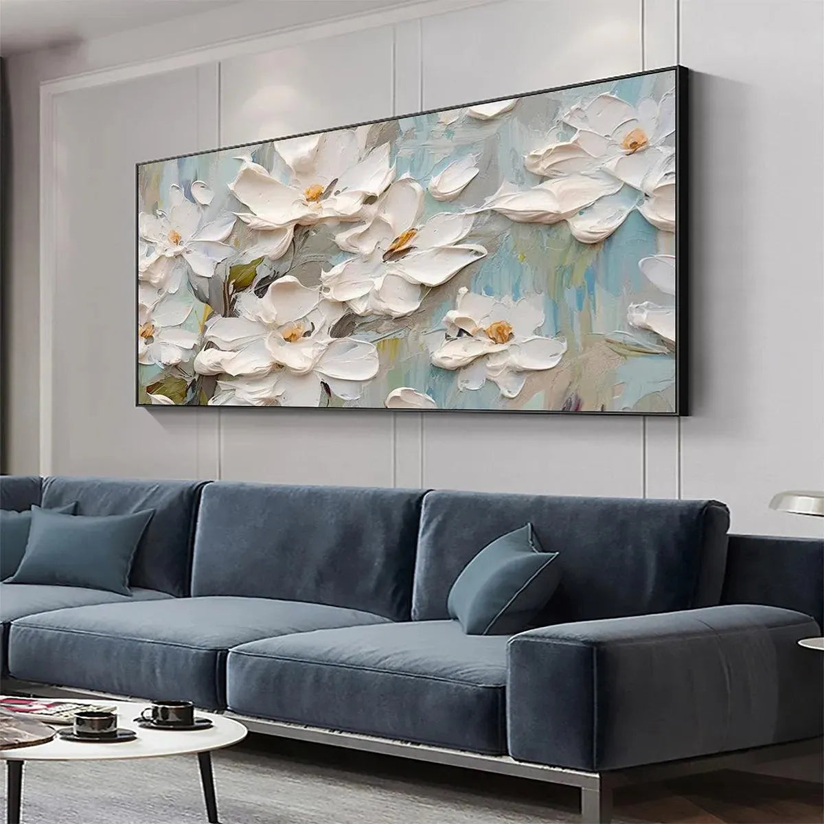 Panoramic white magnolia impasto wall art with textured flowers on soft blue-green canvas, hanging above blue modern sofa.