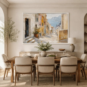 Textured coastal town artwork with visible palette knife strokes, set in a white frame, mounted on a light grey wall above a wooden dining table and chairs.