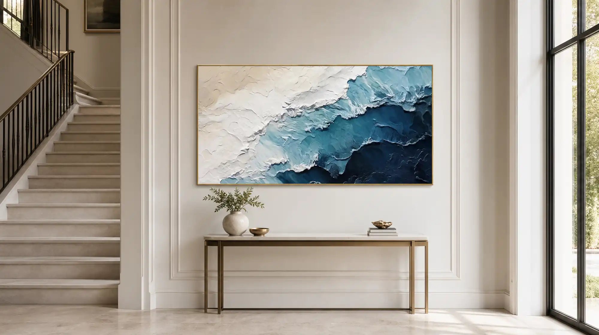 Large horizontal abstract oil painting in blue and cream textures hung on a staircase wall above a console table, demonstrating proper wall art sizing for tall stairwell spaces.